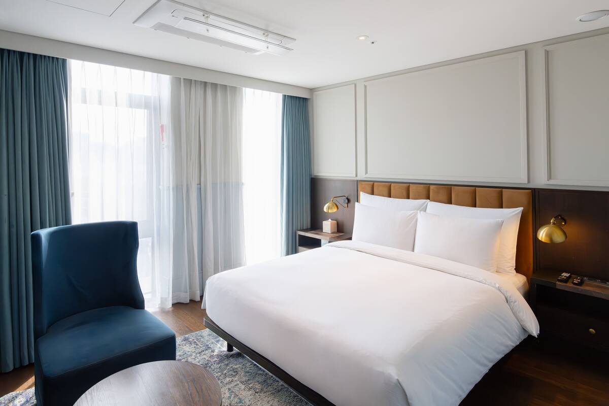 A hotel room with a large bed, a chair, a bedside table with a lamp, and a window with curtains.