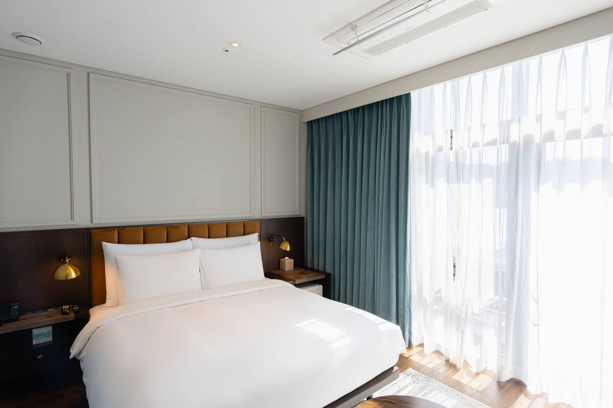 A modern hotel room with a large bed, wooden headboard, bedside lamps, and a view of the outdoors through sheer curtains.
