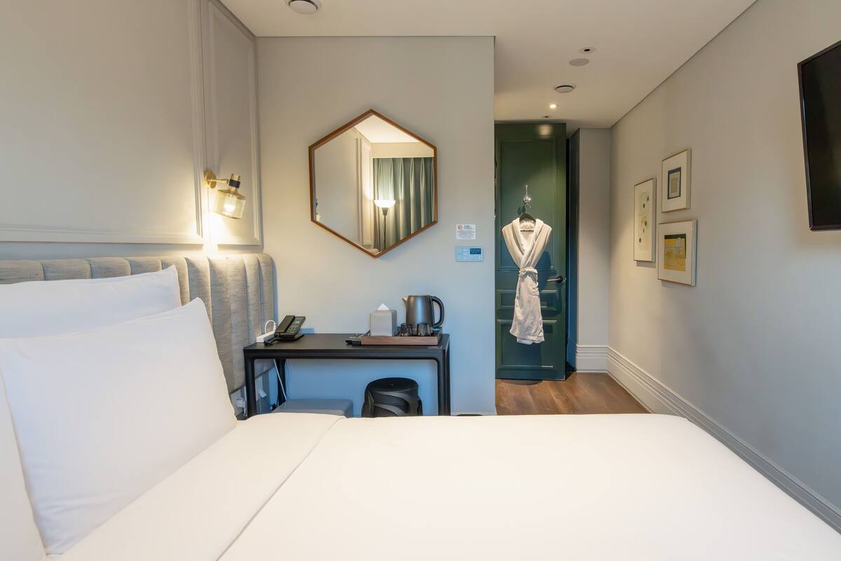 A hotel room with a bed, a desk, a mirror, a robe hanging on the door, and framed pictures on the wall.