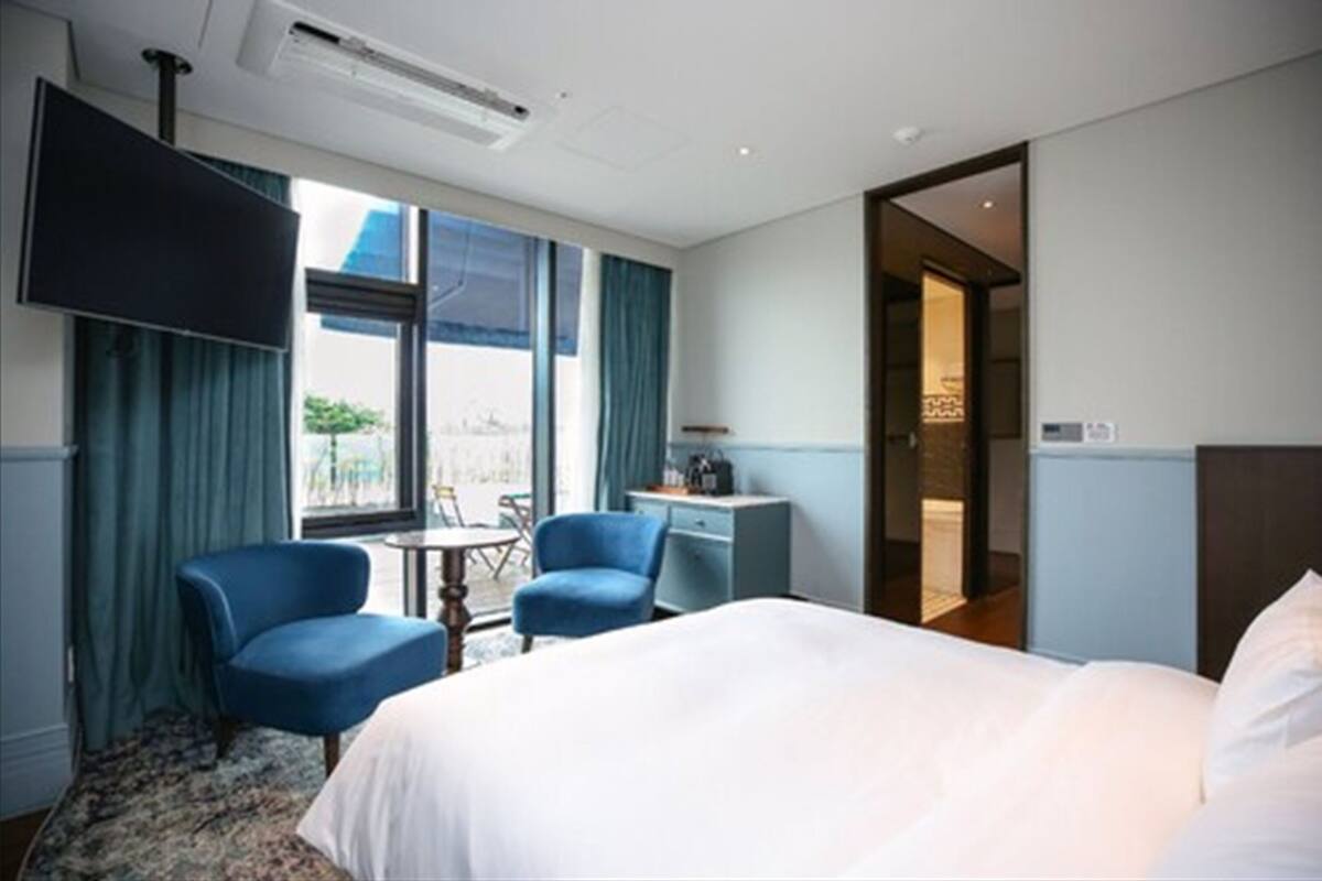 A hotel room with a bed, two blue armchairs, a small table, a desk, and a TV mounted on the wall.