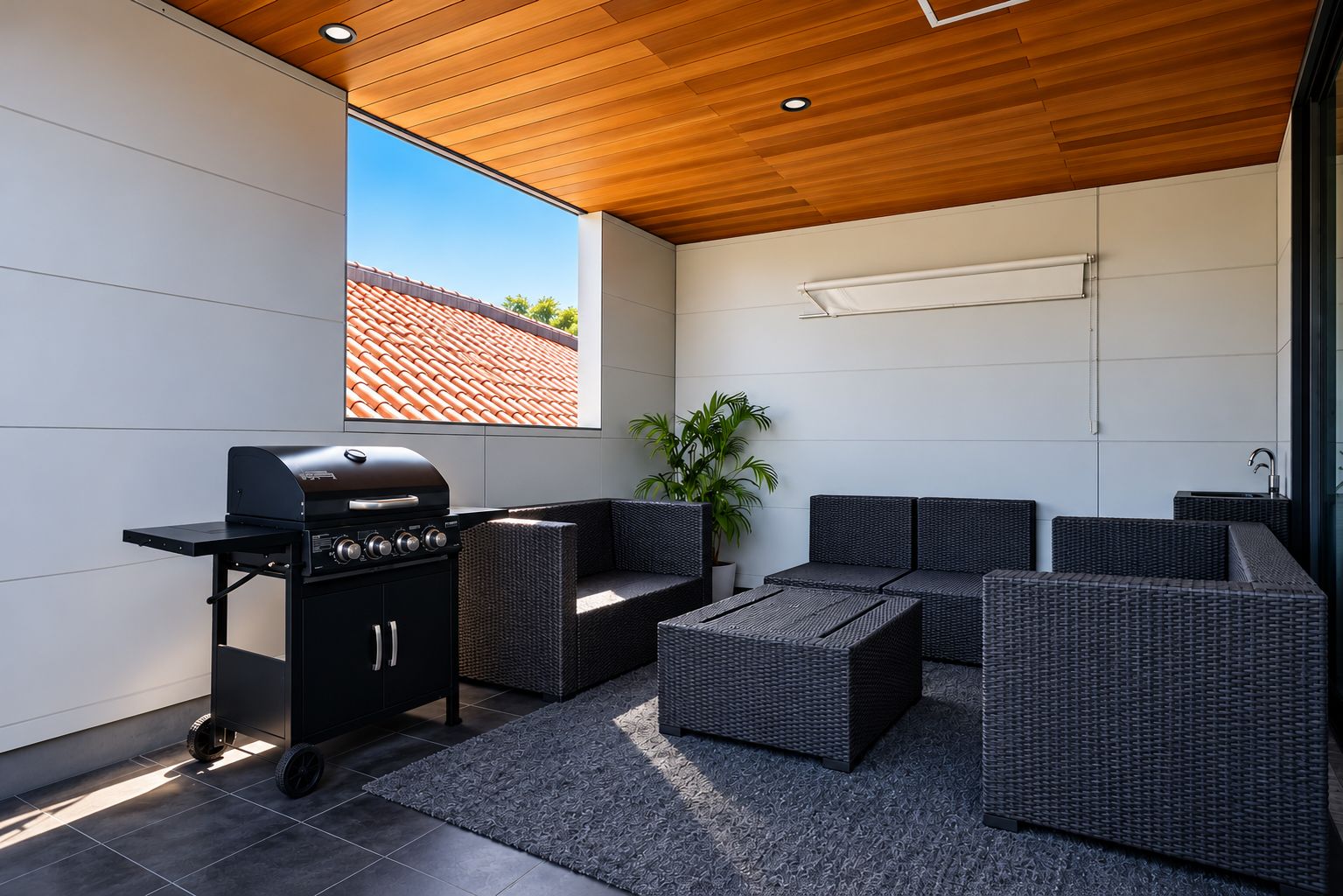 A modern outdoor patio with a grill, wicker furniture, and a view of a red-tiled roof.