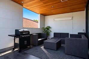 A modern outdoor patio with a grill, wicker furniture, and a view of a red-tiled roof. - TOTONOI Villa YAE (Motobu)