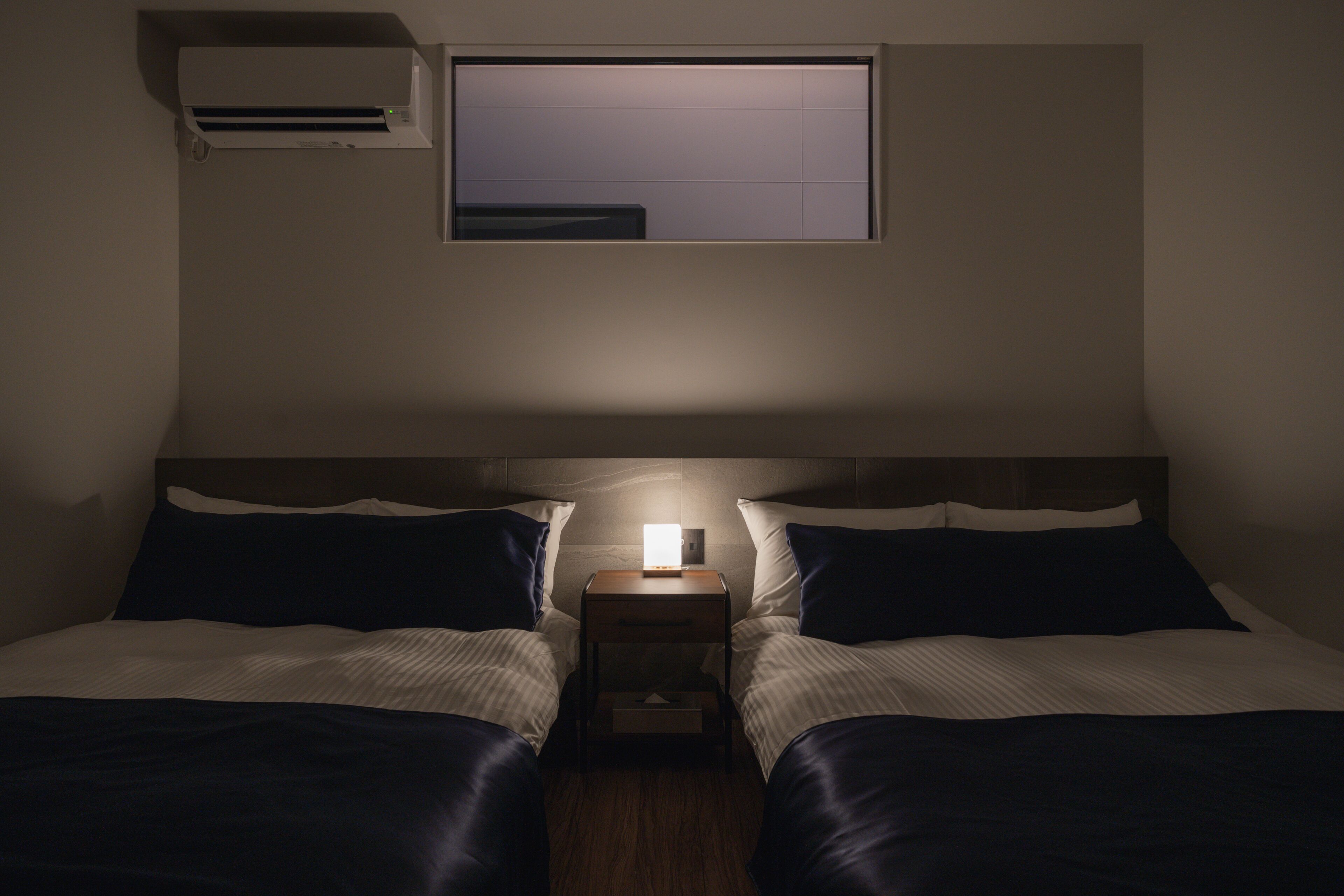 A twin room with two beds, a nightstand, a wall-mounted air conditioner, and a window.
