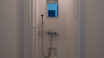Un petit espace douche fermé, équipé d’un pommeau de douche à main, d’un miroir fixé au mur et d’une étagère pour les articles de toilette.