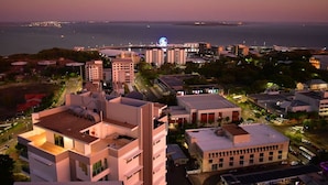 A coastal cityscape at dusk with modern buildings, a prominent Ferris wheel, and a view of the sea.