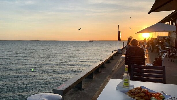A seaside restaurant with outdoor seating overlooking the ocean, a table with a meal, and a sunset in the background.