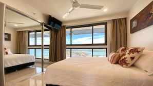 A bedroom with a large bed, a ceiling fan, a TV, and a view of the sea.