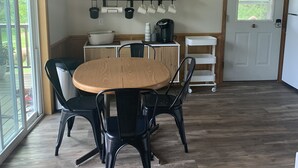 Een moderne keuken met een ronde houten tafel, zwarte stoelen en een hangende lamp. Boven de aanrechttafel staan planken met keukengerei en een deur met een raam.