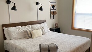 Een net opgemaakt bed met een bedsprei en kussens, twee wandlampen, een hangend wandtapijt en een raam met jaloezieën.