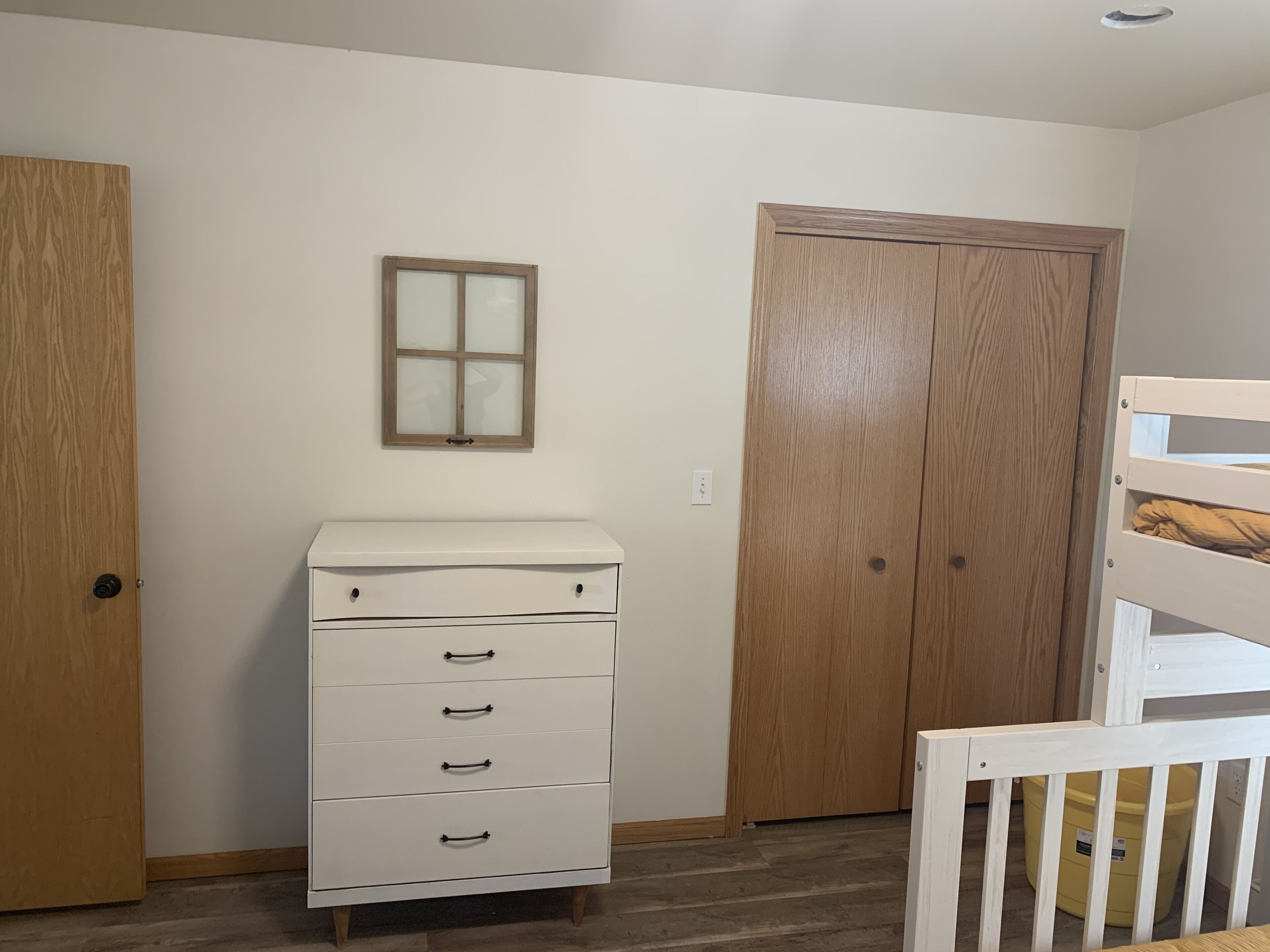 Une pièce avec une armoire en bois, une commode blanche à tiroirs, une porte en bois et une fenêtre à vitres dépolies.