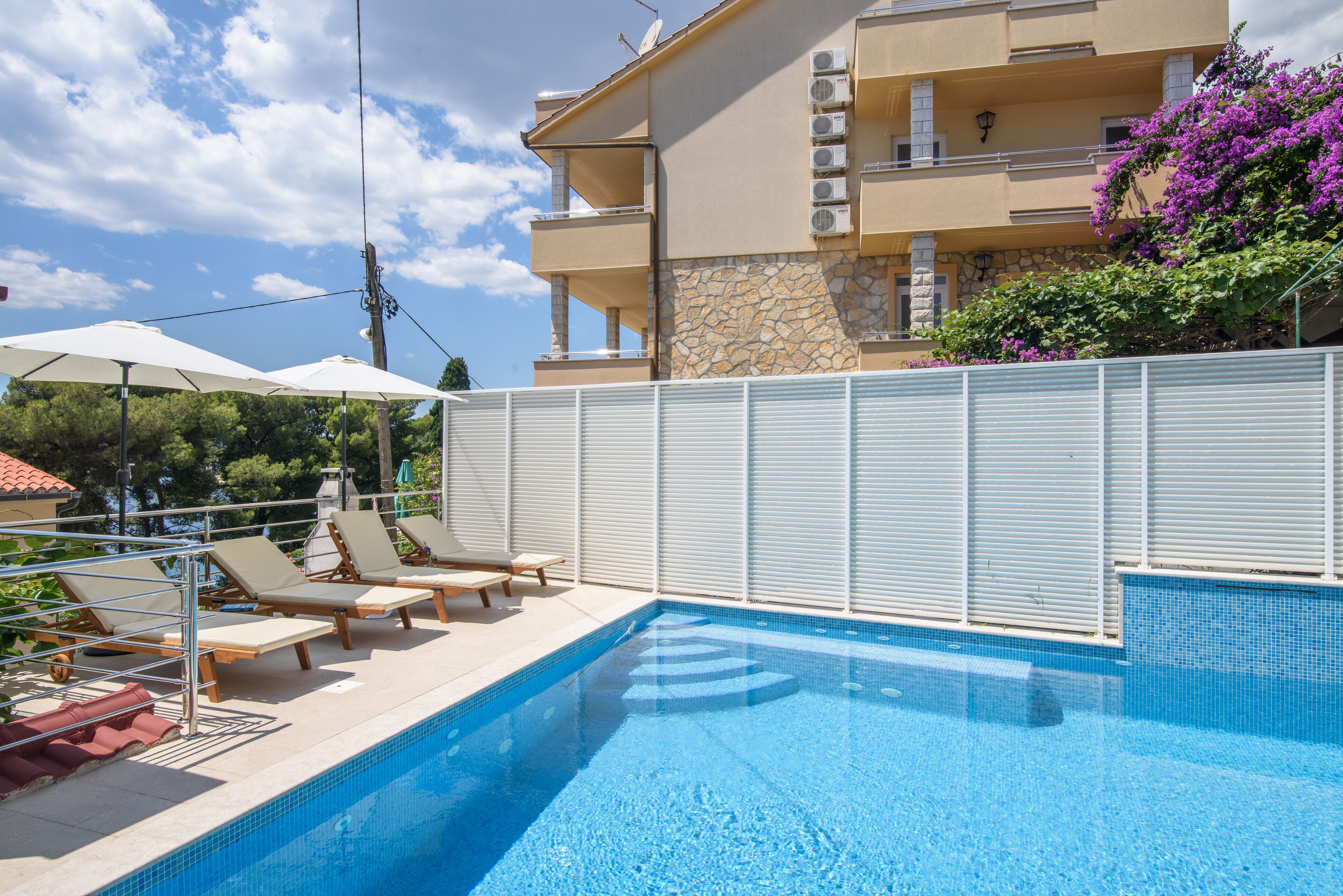 Une piscine entourée de chaises longues et de parasols, le tout délimité par une clôture blanche, avec un bâtiment visible au loin.