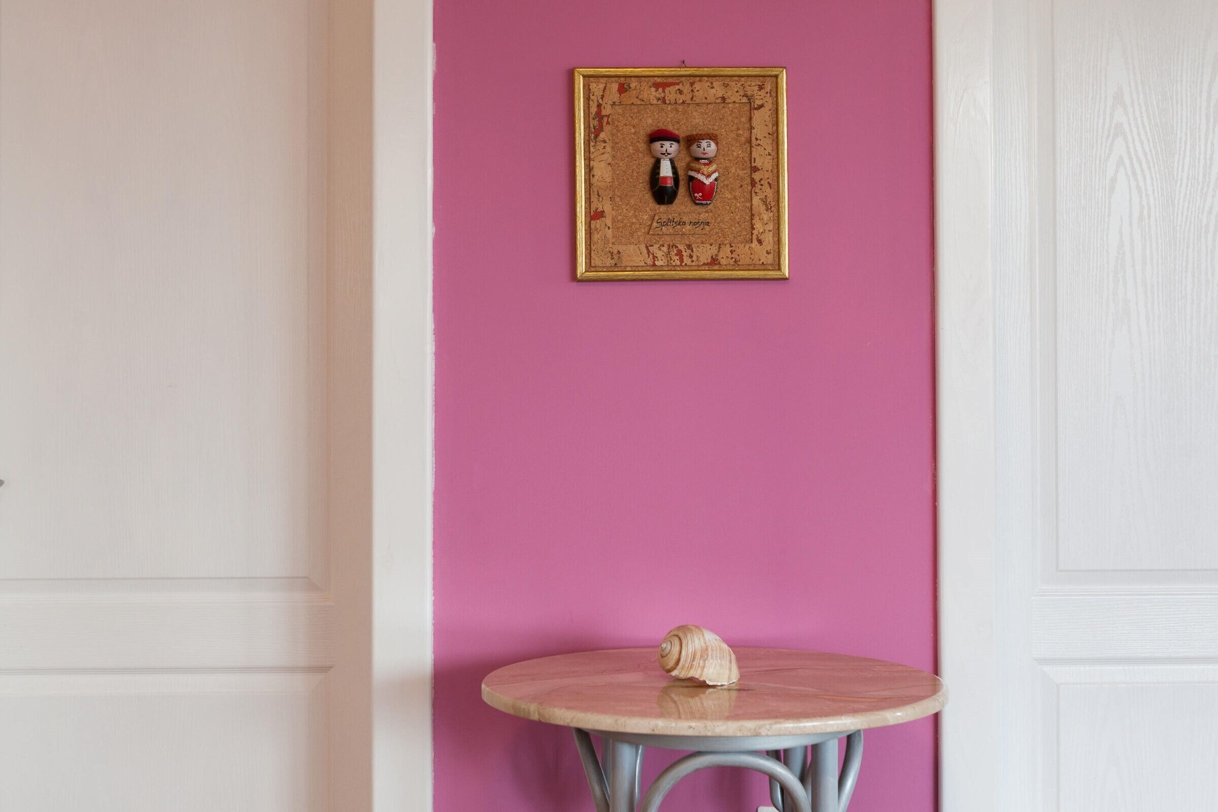 A room with a pink wall, a framed picture, and a round table with a shell on it.
