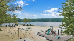 A playground with swings and a slide, located near a lake with a sandy beach.