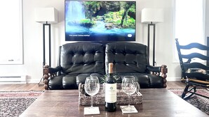 A living room with a black leather sofa, a wooden coffee table with a bottle of wine and two glasses, a flat-screen TV displaying a nature scene, and a patterned rug.