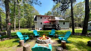 A cabin with a deck, surrounded by trees, and a fire pit with chairs.