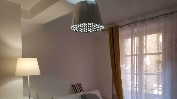 Une chambre à coucher moderne avec un canapé blanc, une table de chevet, une lampe et une fenêtre ornée de rideaux légers.