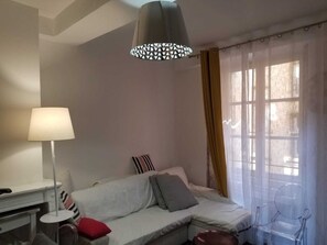 Een moderne slaapkamer met een witte bank, een nachtkastje, een lamp en een raam met dunne gordijnen.