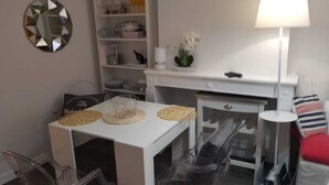 Un espace de vie compact comprenant une table à manger blanche, des chaises en plastique transparent, un meuble blanc avec des étagères, un miroir décoratif, une lampe et une petite plante en pot.