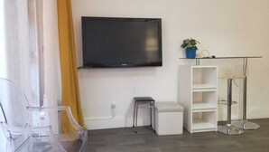 Un salon moderne avec un téléviseur à écran plat fixé au mur, un petit pouf blanc, une table basse en verre et une étagère blanche sur laquelle est posée une plante en pot.