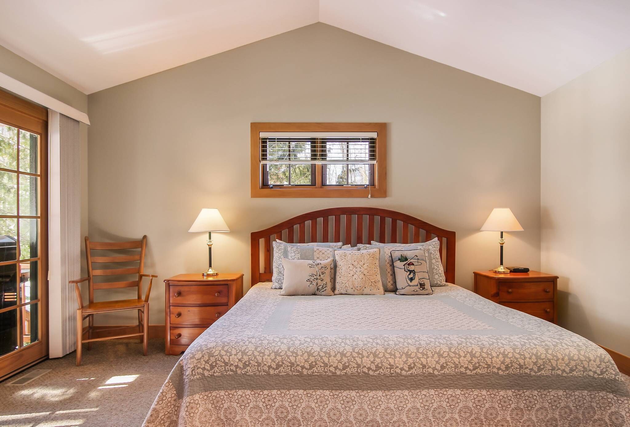 A bedroom with a large bed, two bedside tables with lamps, a wooden chair, and a window with blinds.