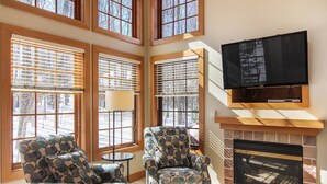 A living room with two patterned armchairs, a fireplace, a TV mounted above it, and large windows with blinds. - Disciples Village beautiful Condo - Steps to ski and lakes (Boyne Falls)