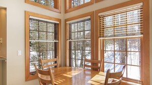 A dining area with a wooden table and chairs, large windows with blinds, and a view of trees outside. - Disciples Village beautiful Condo - Steps to ski and lakes (Boyne Falls)