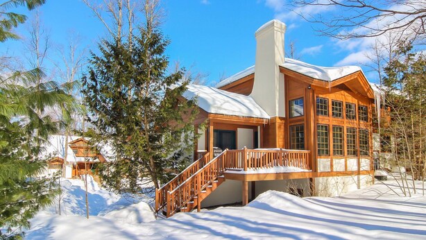 A wooden cabin with a snow-covered roof, a wooden staircase, and a large window. - Disciples Village beautiful Condo - Steps to ski and lakes (Boyne Falls)