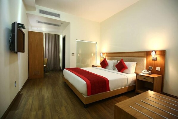 A hotel room with a large bed, a bedside table with a lamp, a mirror, and a wooden floor. - PI Suites (New Delhi)