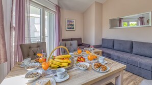 Une table dressée pour le petit-déjeuner, avec une variété d’aliments et de boissons, un canapé gris et une fenêtre avec des rideaux.