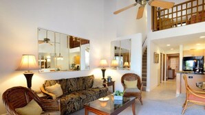 A spacious living area with a sofa, two wicker chairs, a glass coffee table, and a dining table. There are multiple lamps and a ceiling fan. A staircase leads to an upper level. - Ocean-View Maui Kamaole Escape – A/C, Lanai, Walk to Beach & Pool! (Kihei)