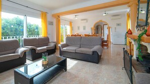 A spacious living area with a sofa set, a glass-top coffee table, and a dining area in the background.