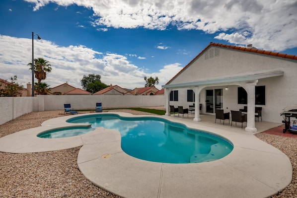 A swimming pool with a patio area, a covered outdoor seating area, and a white building with a red roof.