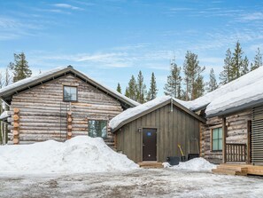 A log cabin with a wooden door and a window, surrounded by snow and trees.