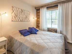 Quarto com cama, mesa de cabeceira, luminária na parede, janela com cortinas e parede de madeira.