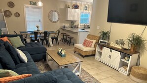 A living room with a dark blue sofa, a wooden coffee table, a wicker chair, a dining area with a table and chairs, a kitchen area with cabinets and a sink, and a flat-screen TV mounted on the wall. - 3 Bedroom house in Purple Parrot (Pensacola)