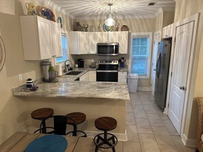 A kitchen with white cabinets, a marble countertop, a built-in microwave, a refrigerator, and a sink. There are three stools at the kitchen island.