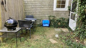 Un jardin avec un barbecue, une table et des chaises. On y trouve une clôture en bois, une fenêtre et quelques plantes.