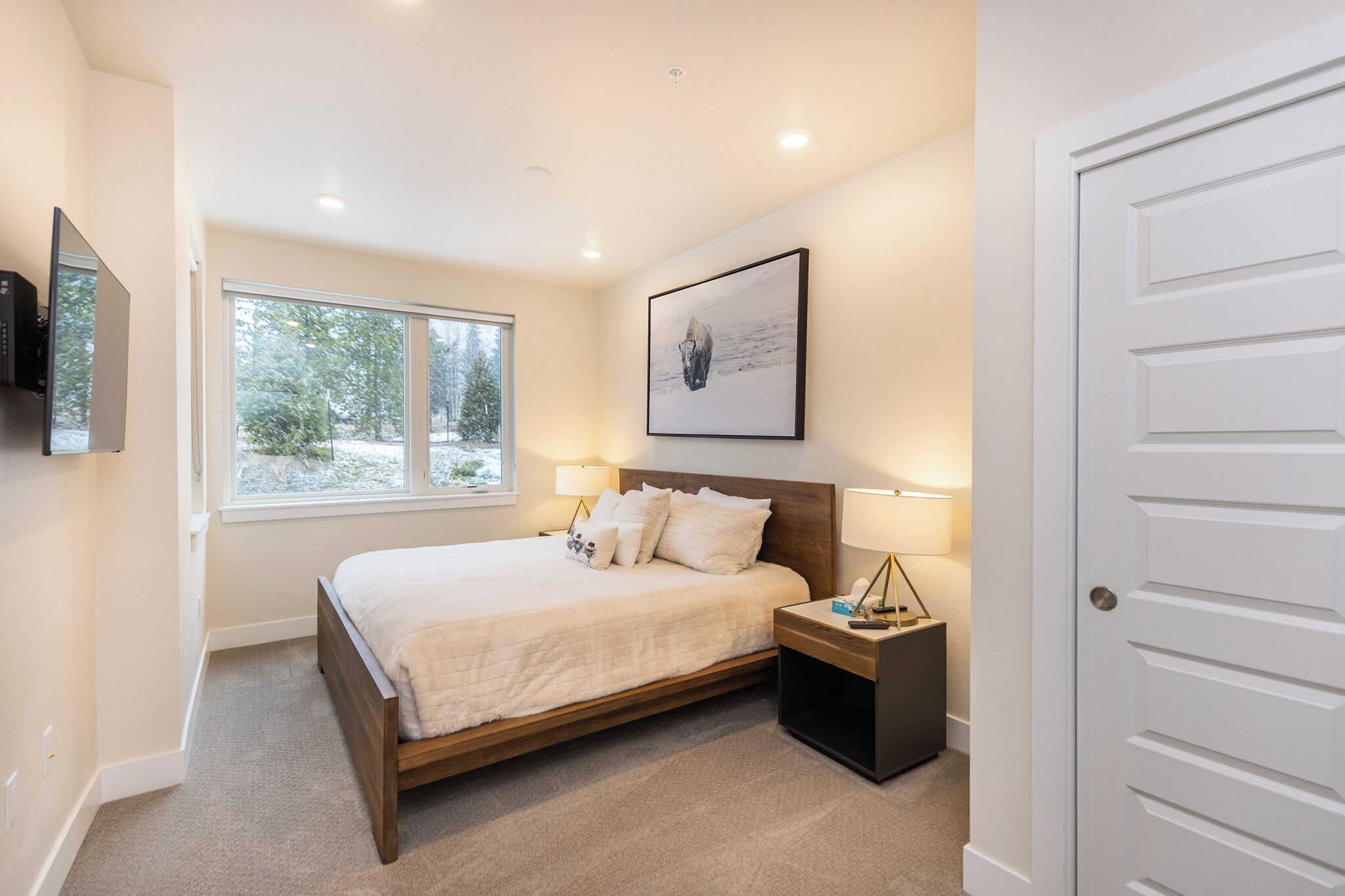 A bedroom with a bed, bedside tables, a TV mounted on the wall, a window with a view of trees, and a door.