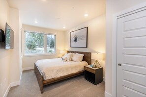 Quarto com cama, luminárias de cabeceira, TV fixada na parede e janela com vista para árvores.