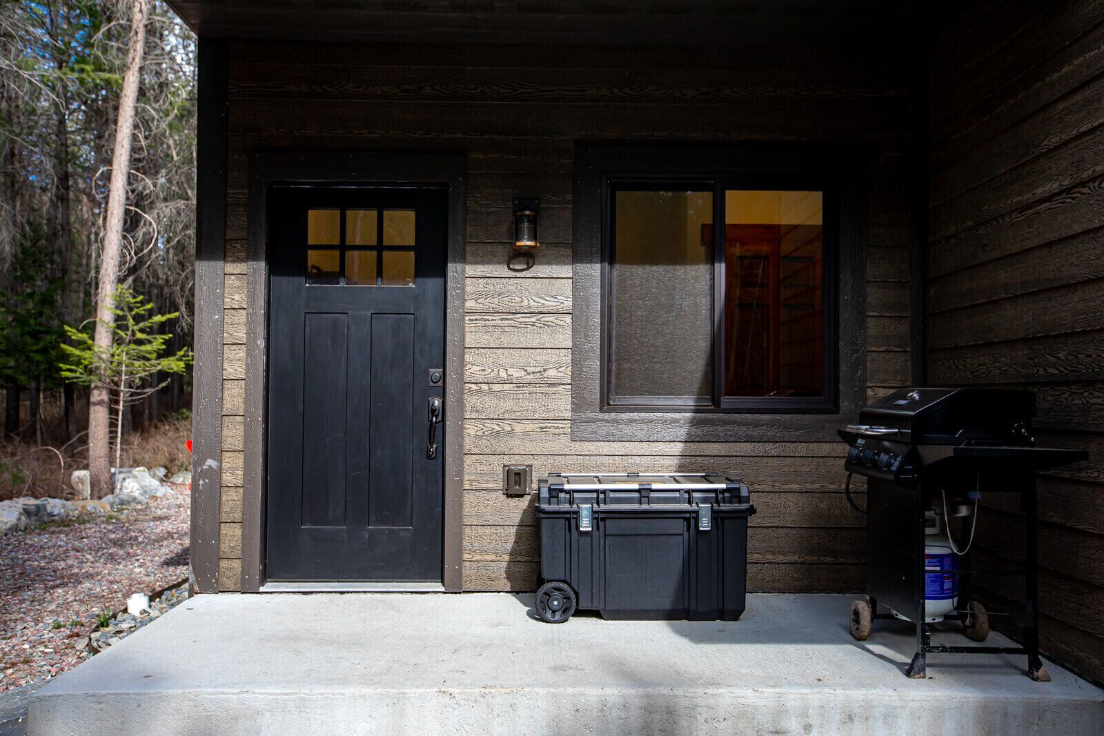 A wooden house with a black door, a window, and a barbecue grill on the porch.