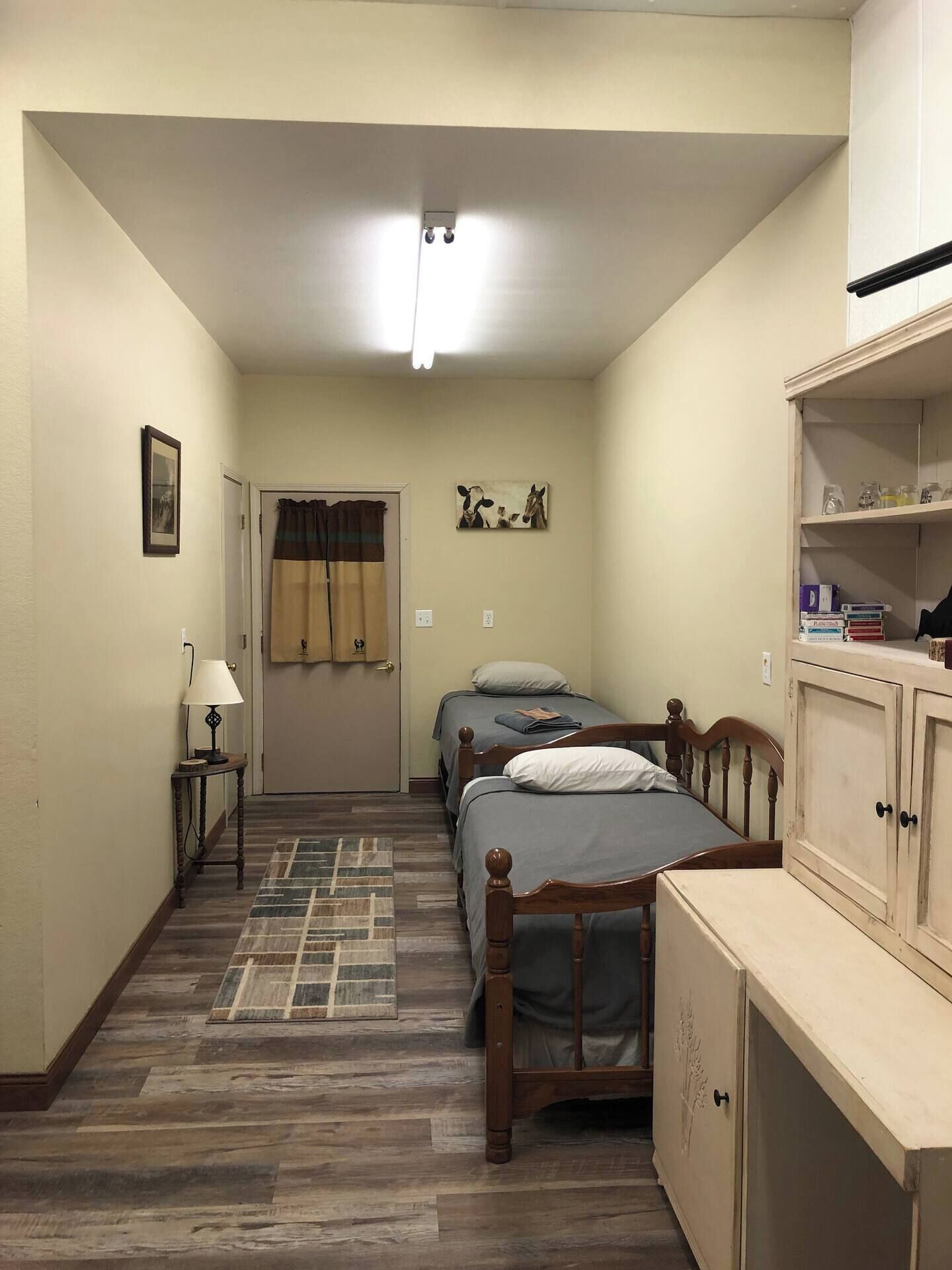 Un pasillo estrecho que conduce a un dormitorio con dos camas, una mesita de noche con una lámpara, un armario y una puerta con cortinas.