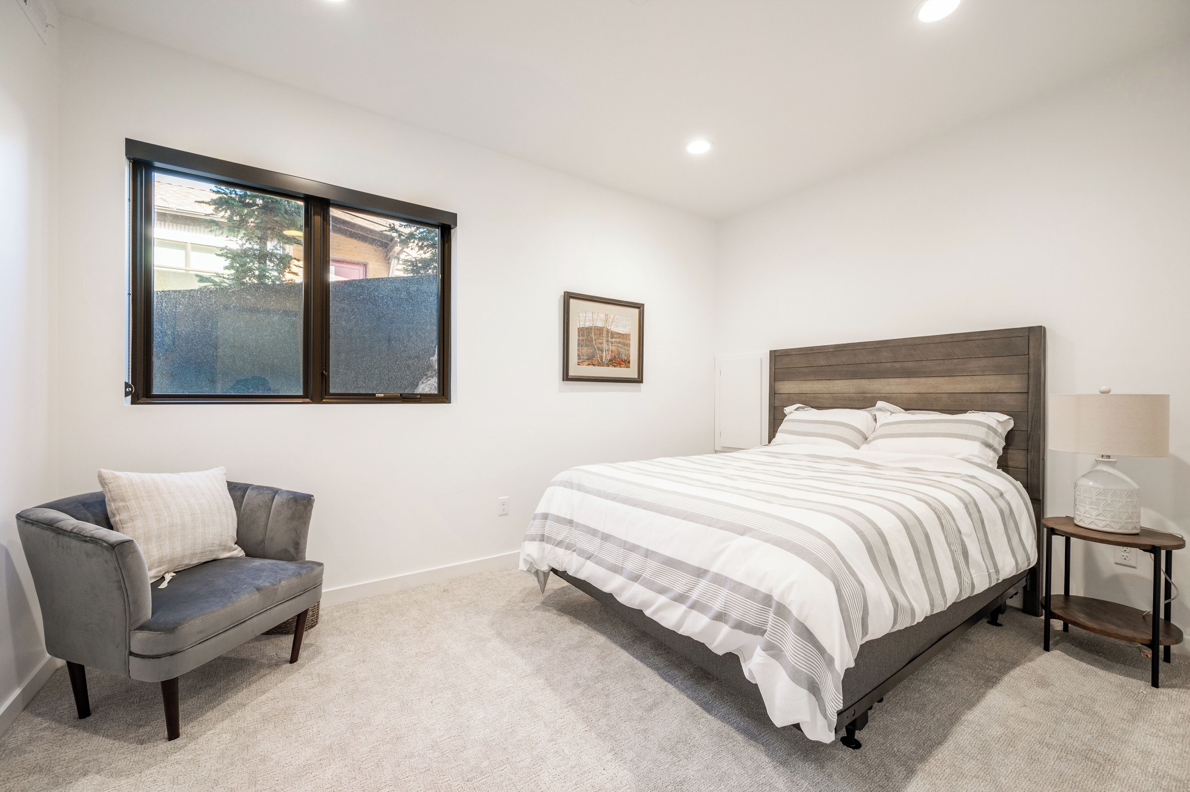A bedroom with a bed, a gray armchair, a nightstand, a window, and a framed picture on the wall.