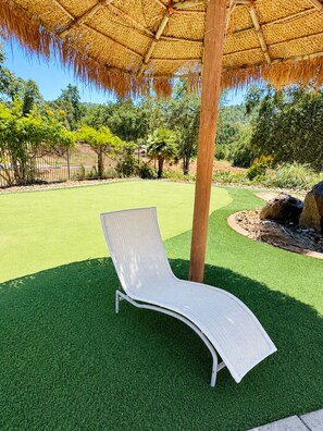 Een lounge stoel onder een rieten parasol in een verzorgde tuin met kunstgras en diverse planten.