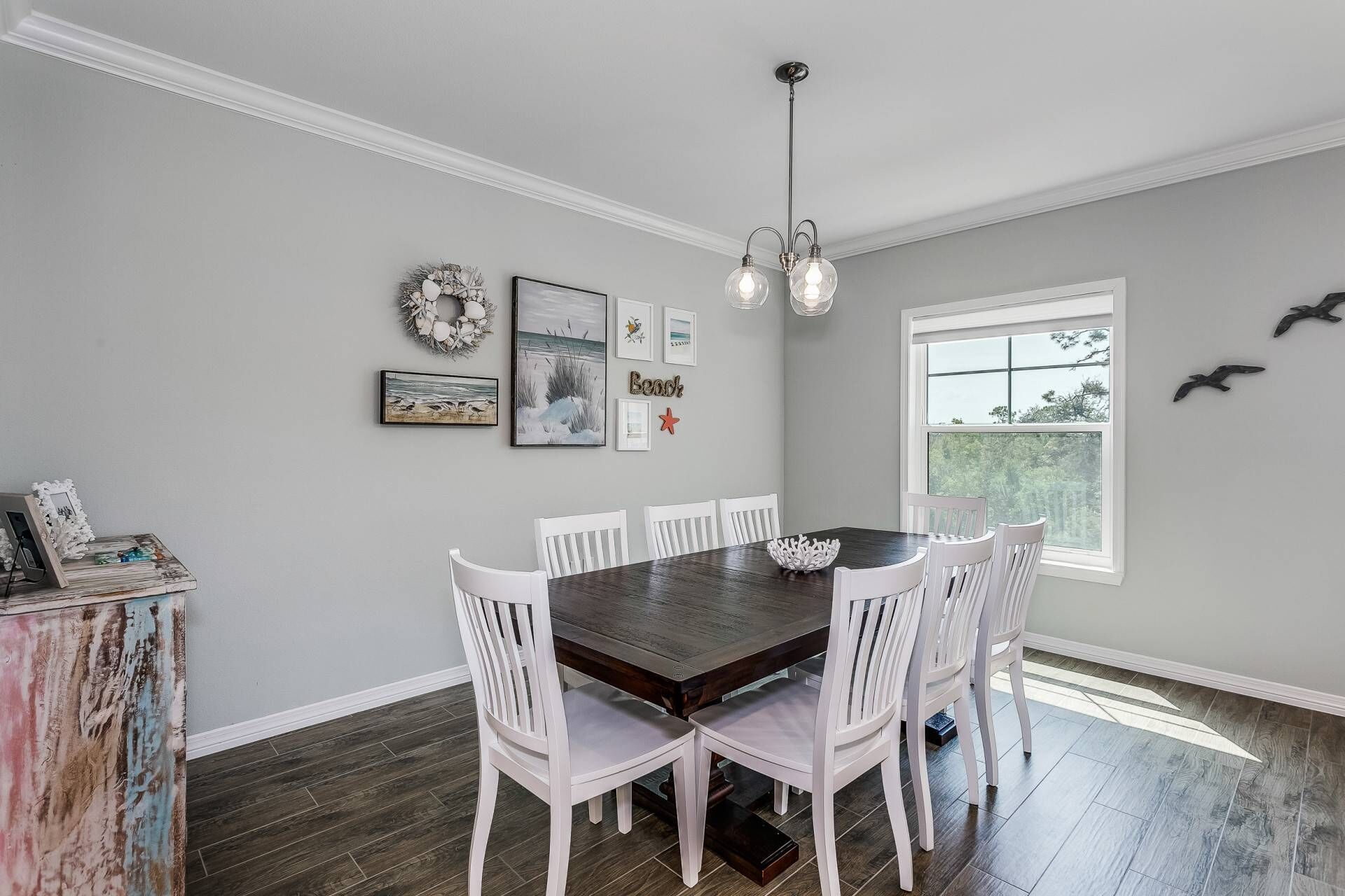 Une salle à manger avec une table en bois foncé, entourée de chaises blanches, un lustre et des éléments de décoration murale, notamment des tableaux encadrés et une étoile de mer.