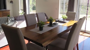 Uma sala de jantar com uma mesa de madeira, quatro cadeiras, duas plantas em vasos, um relógio na parede e um sofá ao fundo.