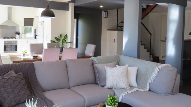 Uma sala de estar moderna com um sofá seccional cinza, uma área de jantar com mesa e cadeiras, e uma cozinha ao fundo.