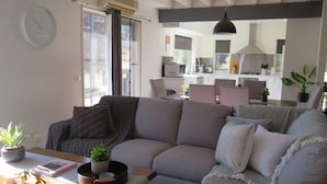 Uma sala de estar moderna com um sofá seccional cinza, uma mesa de centro de madeira e uma área de jantar, com uma cozinha ao fundo.