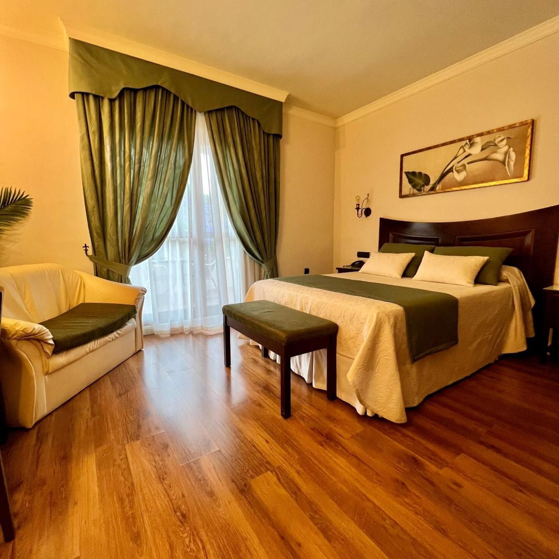 Una habitación de hotel con cama, una silla, un banco, una mesita de noche, una lámpara de pared y un cuadro.