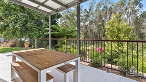 Une terrasse couverte avec une table en bois et des bancs, entourée d’une rambarde en métal et d’une végétation luxuriante.