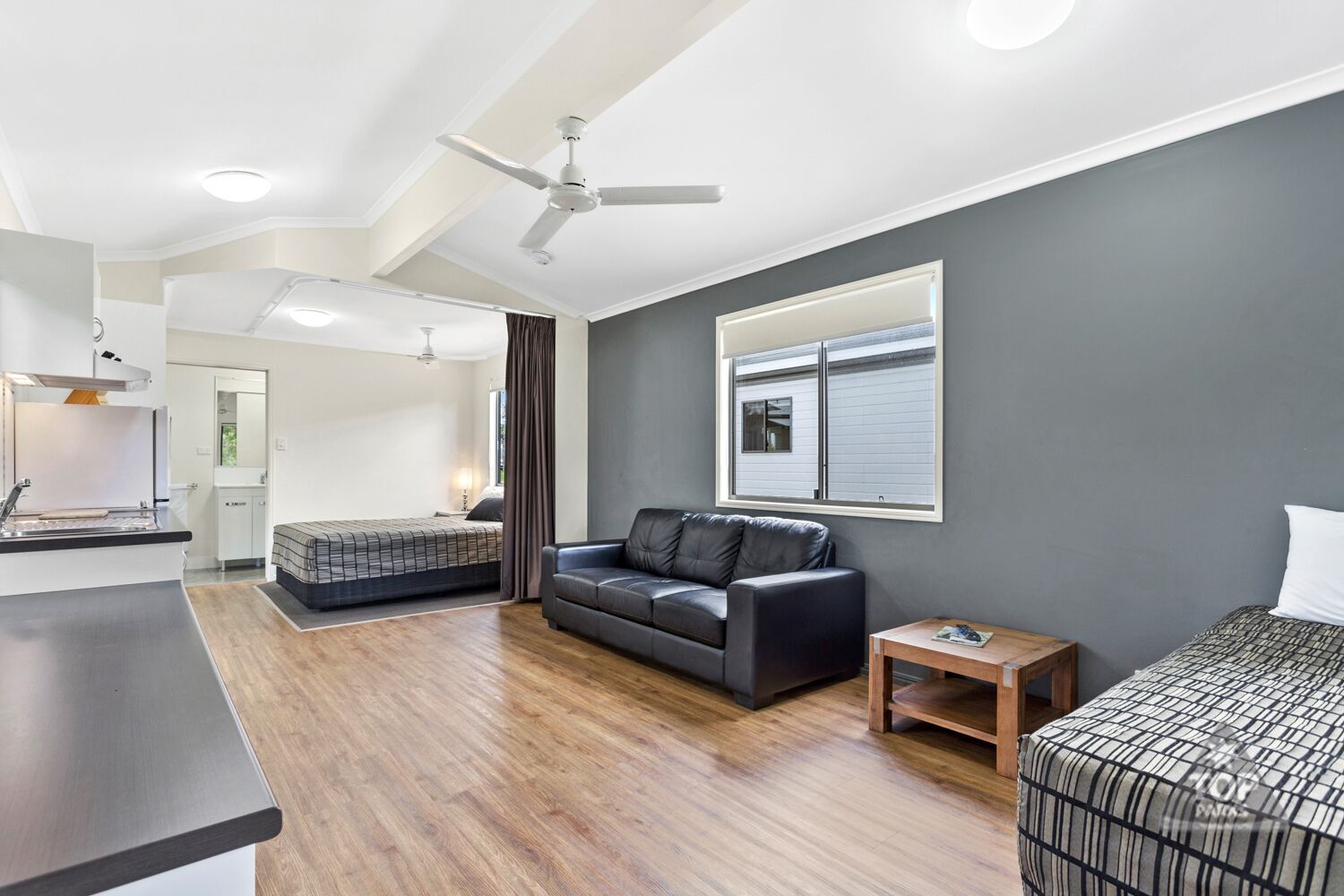 Ein moderner, gut beleuchteter Raum mit Holzboden, einem schwarzen Ledersofa, einem Bett mit gemustertem Überwurf und einem kleinen Holztisch. Ein Deckenventilator ist ebenfalls vorhanden.